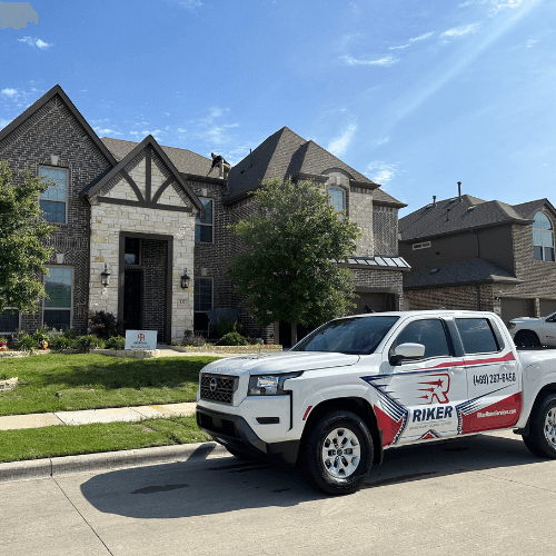 A Riker Home Services work truck is parked in front of a large, modern suburban home with a brick and stone exterior. The truck features the Riker Home Services logo and contact information, and is situated on a well-maintained residential street under a partly cloudy sky.